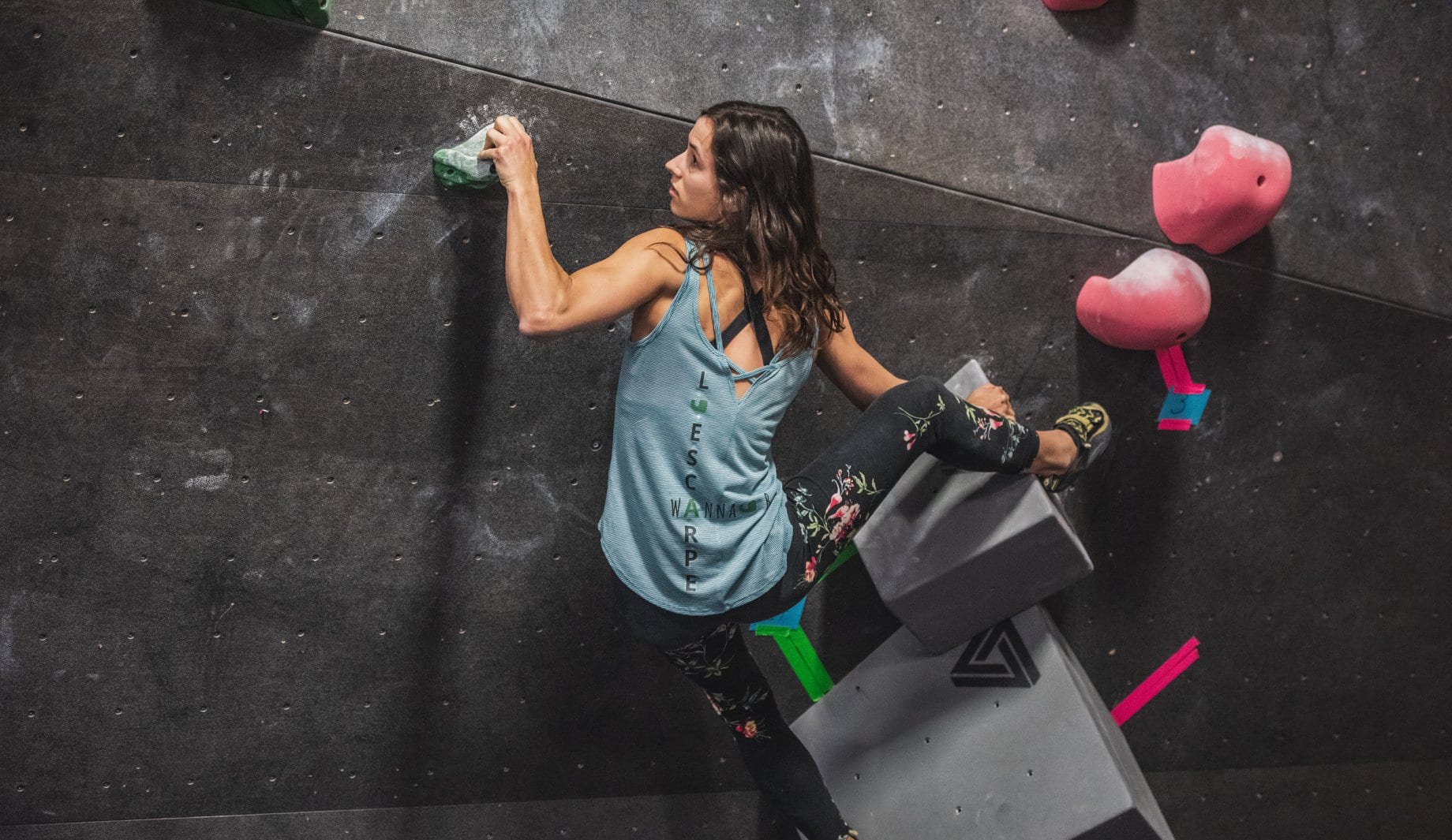 A climber in a teal tank top mid-move on a dark wall